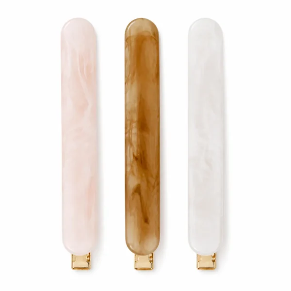 Three acetate French barrettes in the rounded bar profile standing upright on white — pale pink pearl, brown tortoiseshell and white pearl — each on a gold hinged spring bar