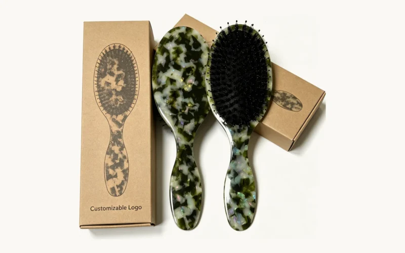 Two acetate cushion hair brushes beside plain kraft boxes marked for customisable logo printing