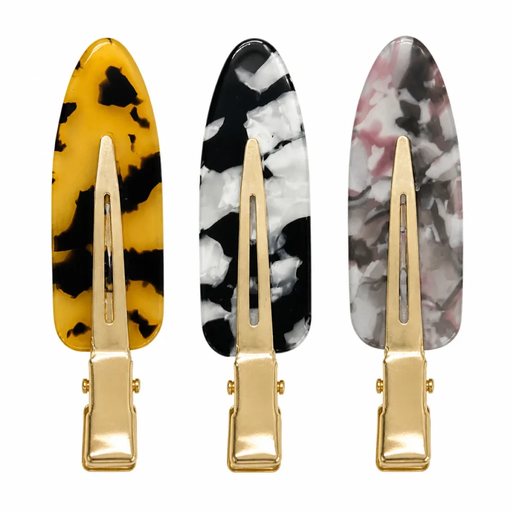 Three acetate duckbill hair clips on gold alligator bases standing upright on white, in yellow tortoiseshell, black-and-white marble and pink-grey marble