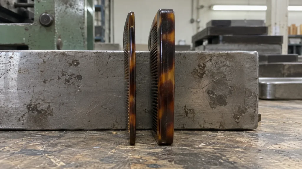 Two combs of the same outline standing on edge against a metal block on a workbench, one visibly thinner than the other