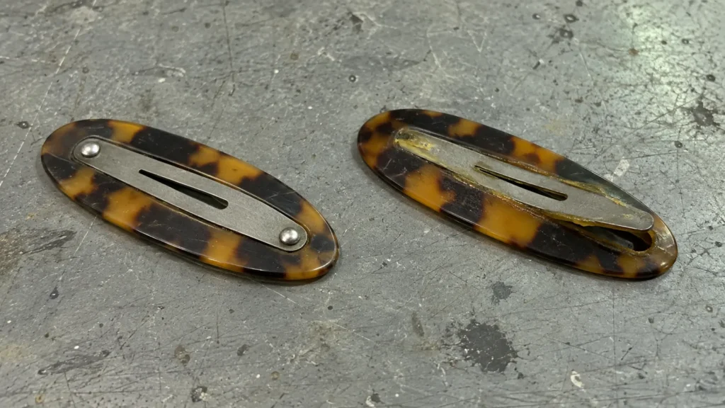 Two tortoiseshell snap clips face down: the left has its steel bar riveted through the acetate, the right has a glued bar lifting at one end